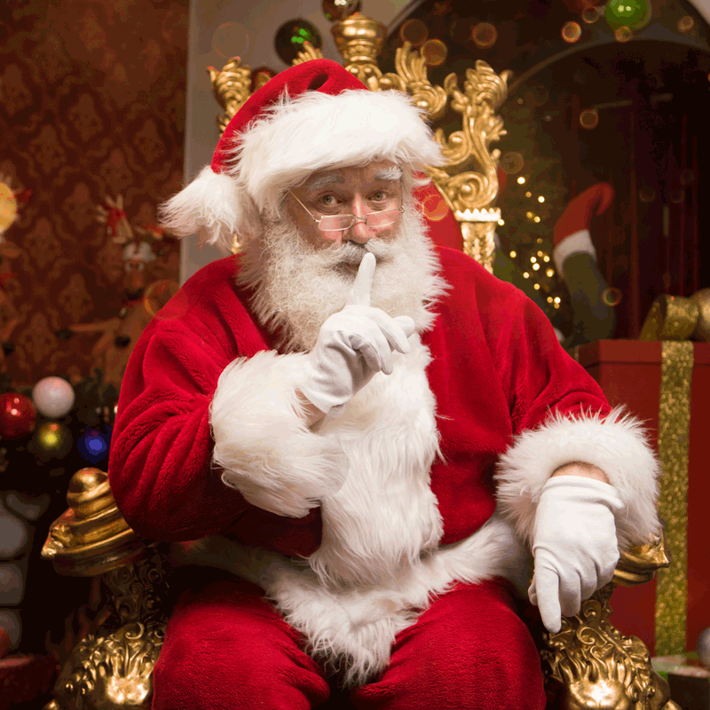 Santa Claus sitting in a decorated Christmas setting, holding a finger to his lips in a shushing gesture.