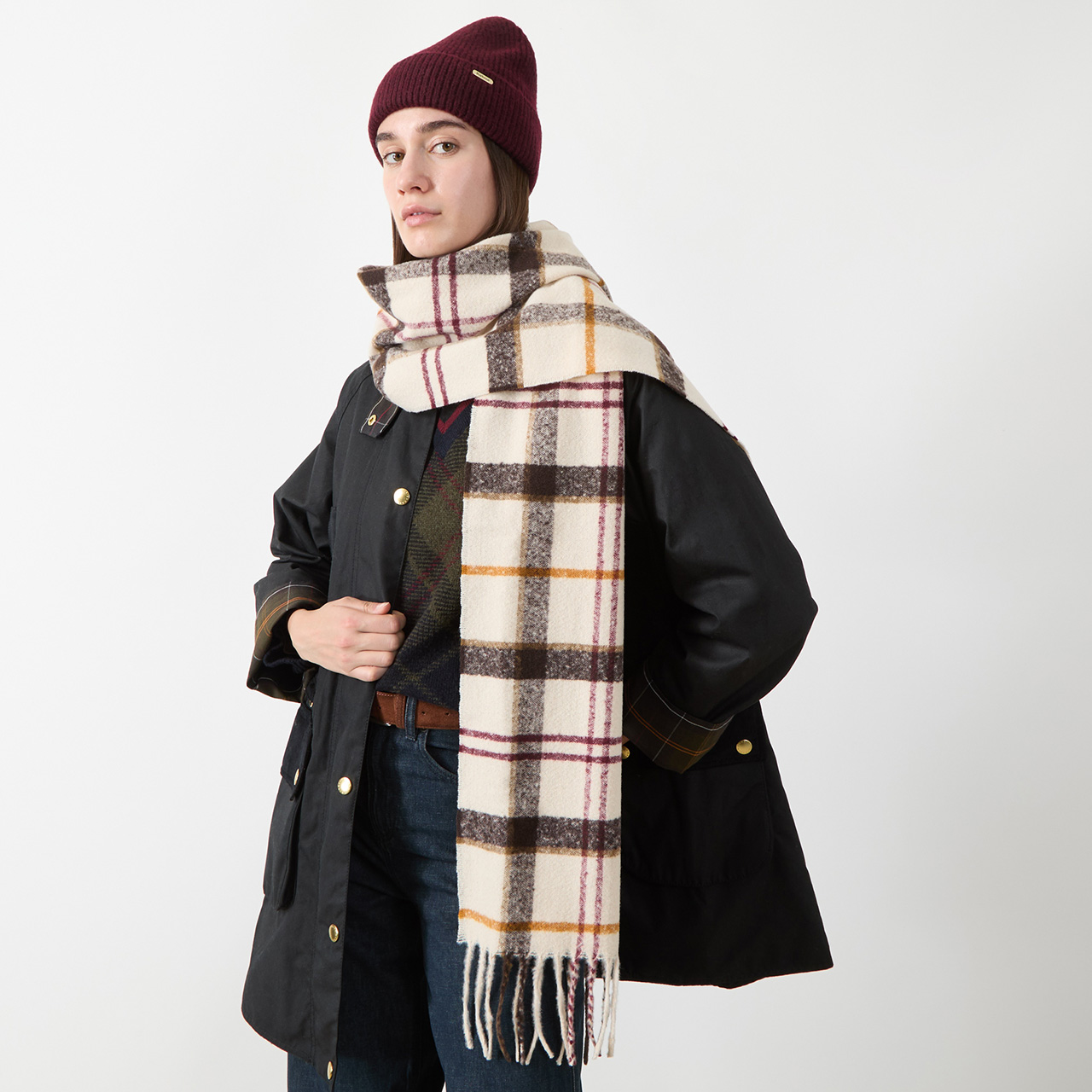 A person wearing a maroon beanie, black coat, and long plaid scarf with fringe, stands against a plain white background. The scarf features broad black and white checks with subtle yellow and red lines. Their hand is on their side.