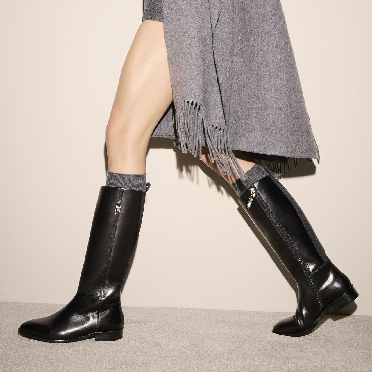 A person wearing a grey fringed coat and black knee-high leather boots with side zips walks against a cream background. Only the legs and lower body are visible.