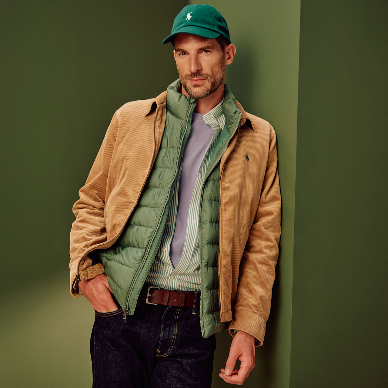 A man with a beard wearing a green baseball cap, beige jacket, green quilted gilet, green striped shirt, and a white T-shirt stands against a green background. He's dressed in dark trousers, leaning casually with his hand in his pocket.