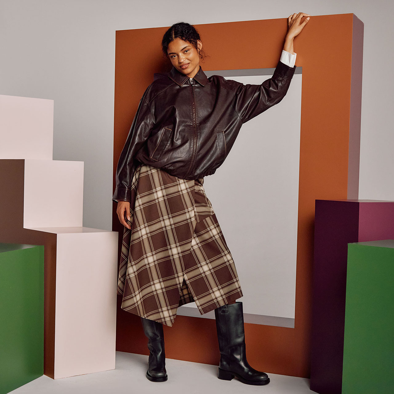 A person stands in a studio setting, wearing a dark leather jacket, a plaid skirt, and knee-high black boots. They lean against an orange frame, with one arm resting on top. Geometric blocks in pink, green, and purple are arranged around them. The background is grey, creating a modern and stylish atmosphere.