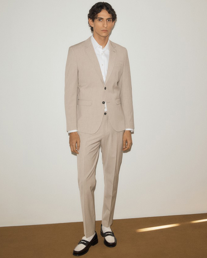 A man in a beige suit and white shirt stands against a plain background, wearing black and white shoes.
