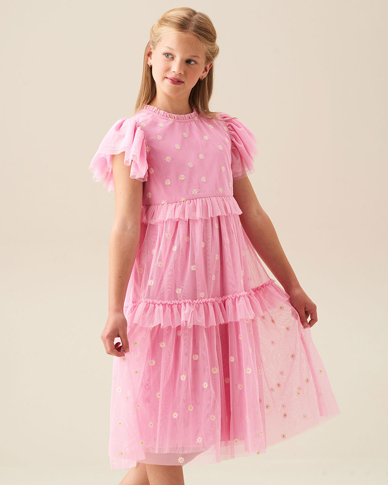 A young girl in a pink, flowy dress with floral patterns and ruffled sleeves looks to the side.

