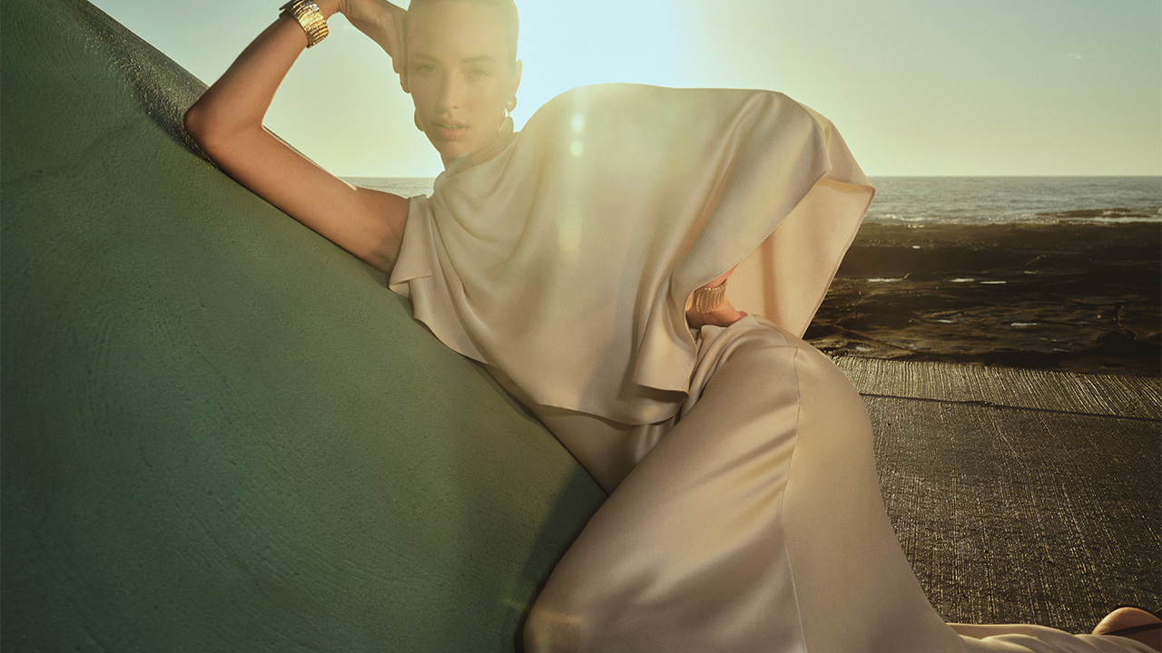 A woman in an elegant cream dress poses outdoors, leaning on a green surface with sunlight in the background.