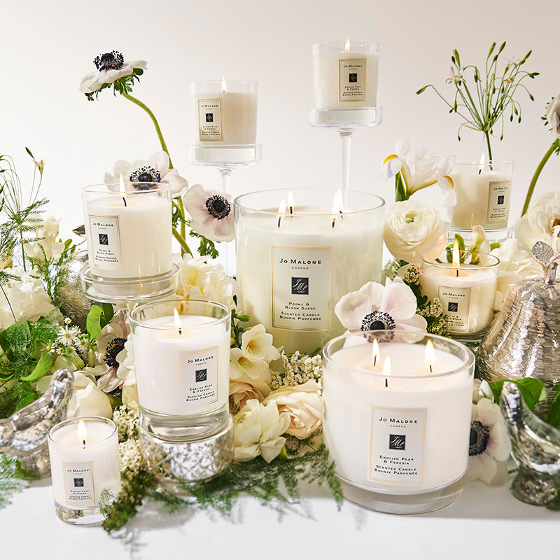A display of Jo Malone candles arranged with white flowers and greenery on a reflective surface.