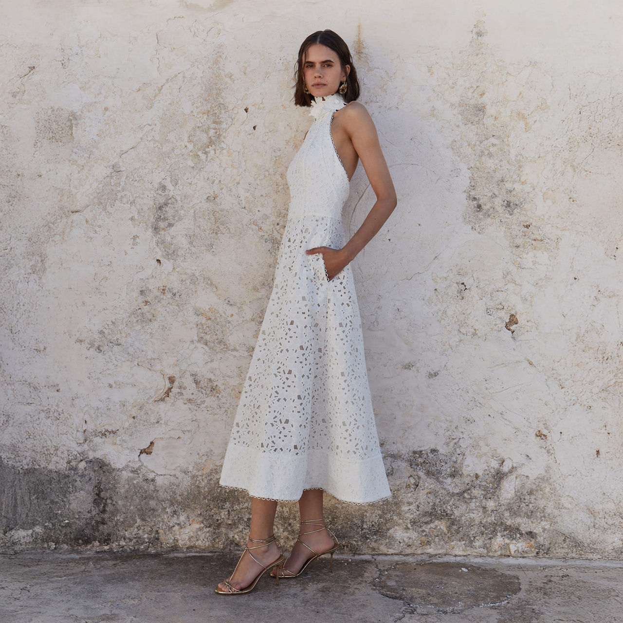 A woman in a sleeveless white dress stands against a textured beige wall.

