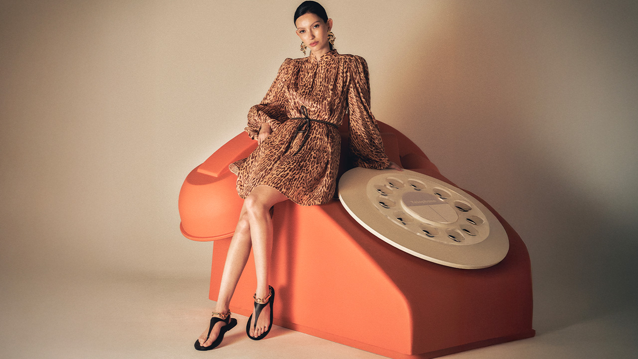 A woman in a patterned dress sits on a giant, vintage-style orange telephone.