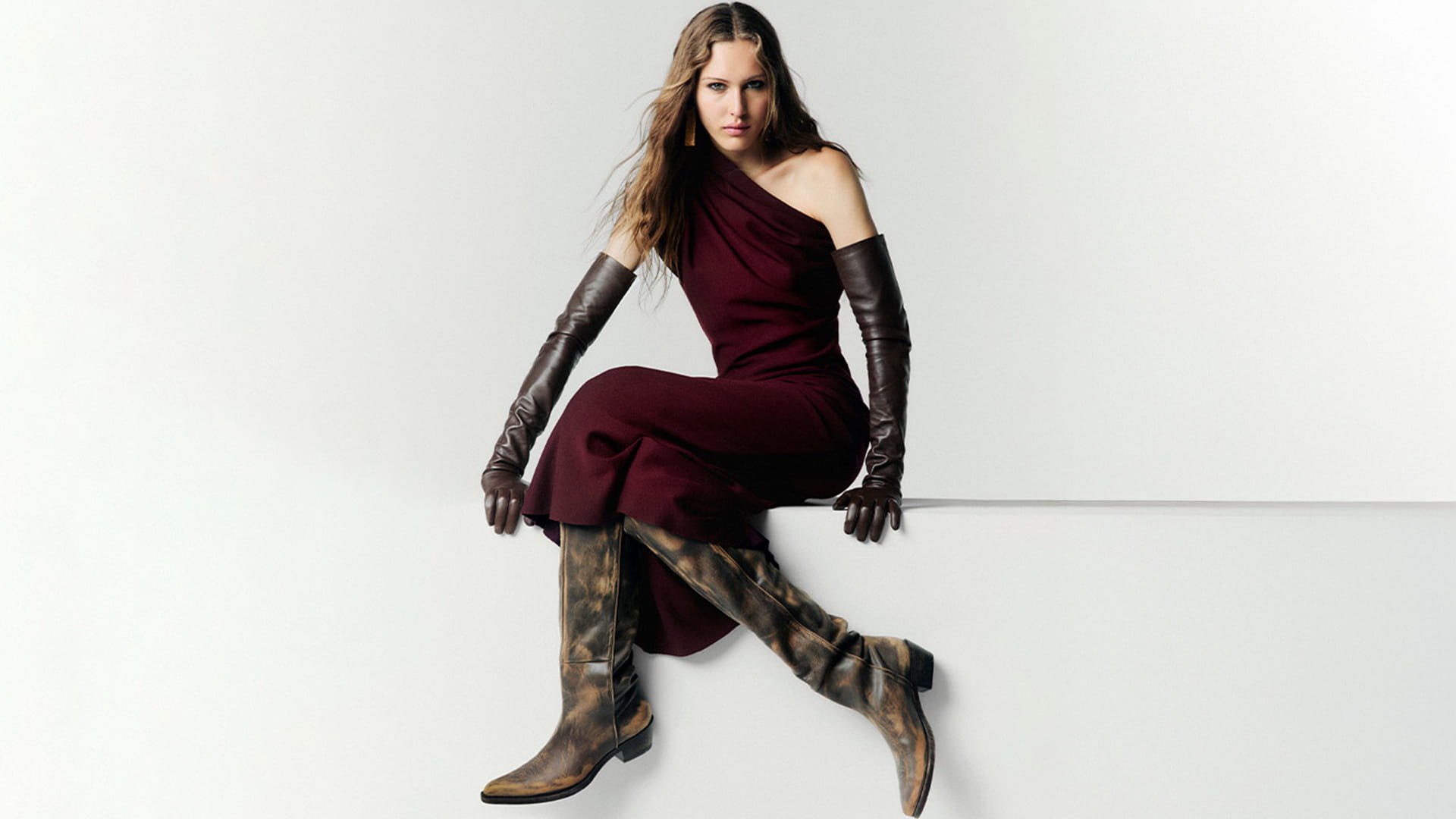 A person sits against a white background wearing a one-shoulder burgundy dress, long gloves, and knee-high patterned boots.