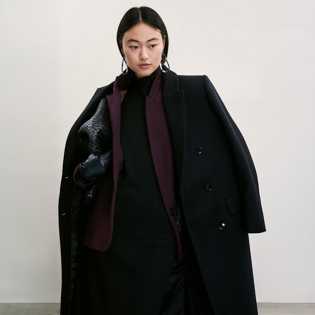 A person wearing a dark coat, burgundy scarf, and large earrings, holding a textured bag.
