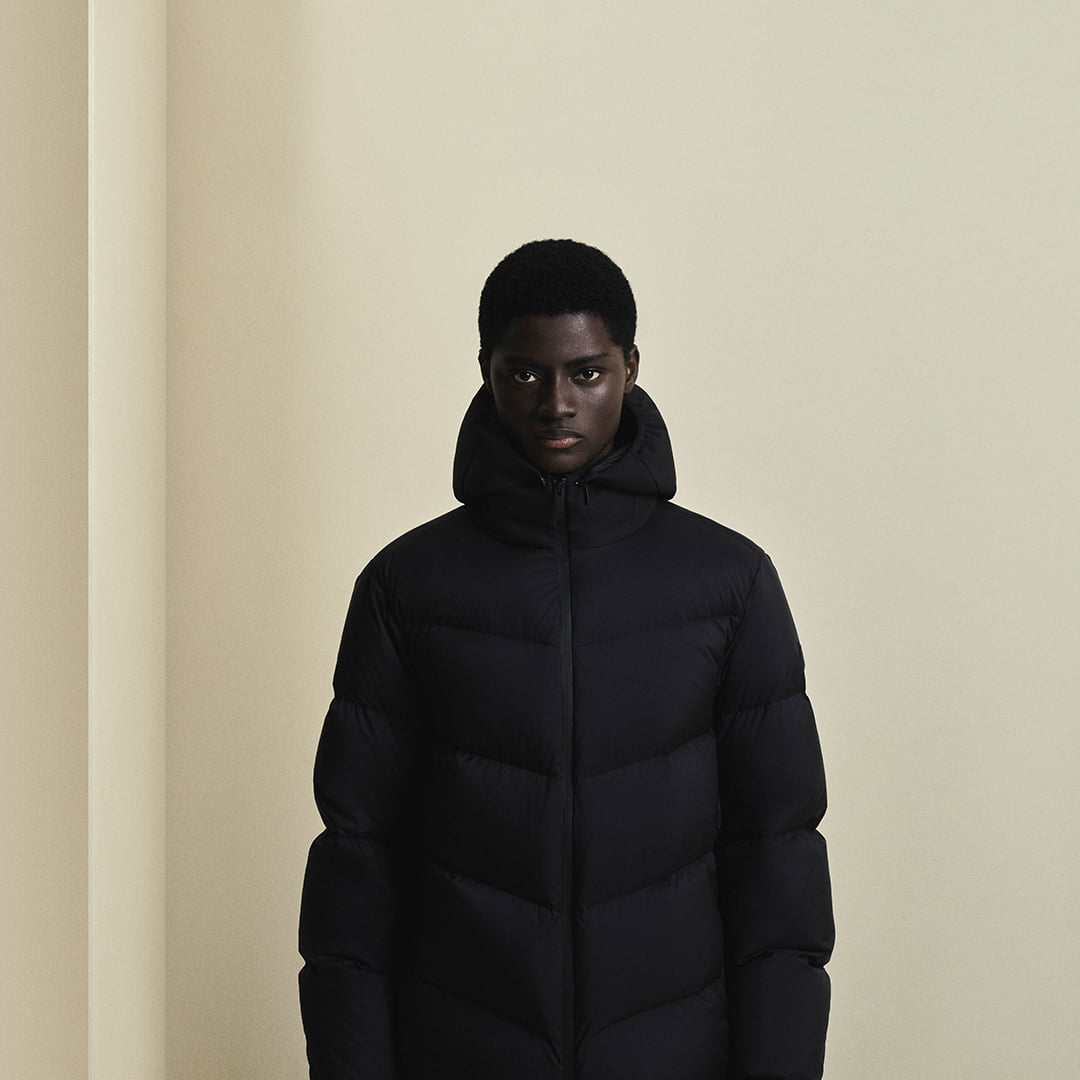 Person wearing a black puffer jacket stands against a beige wall.
