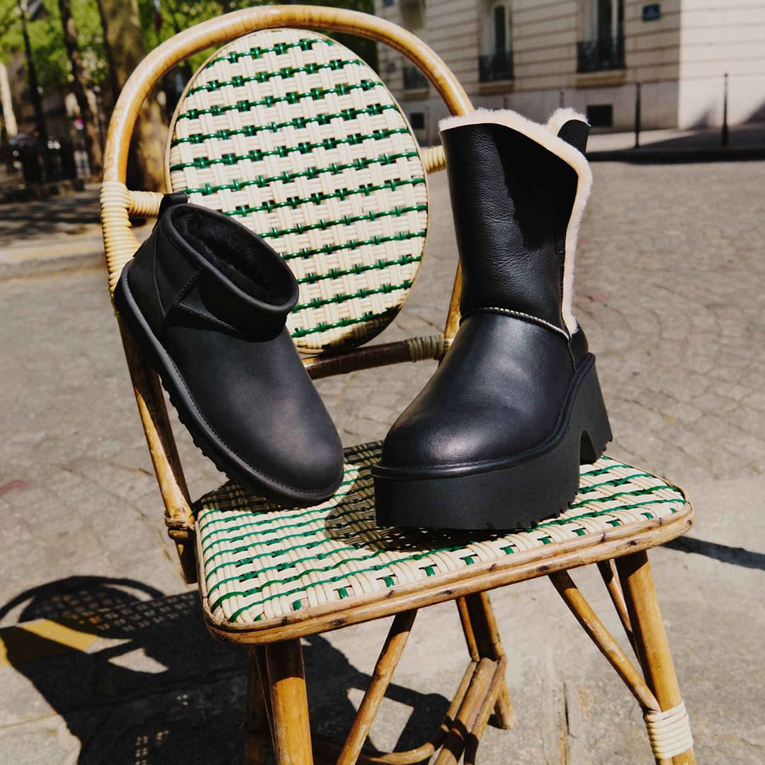 Two black ankle boots with fur lining are displayed on a wicker chair with green and white woven design, set outdoors on a sunny day.
