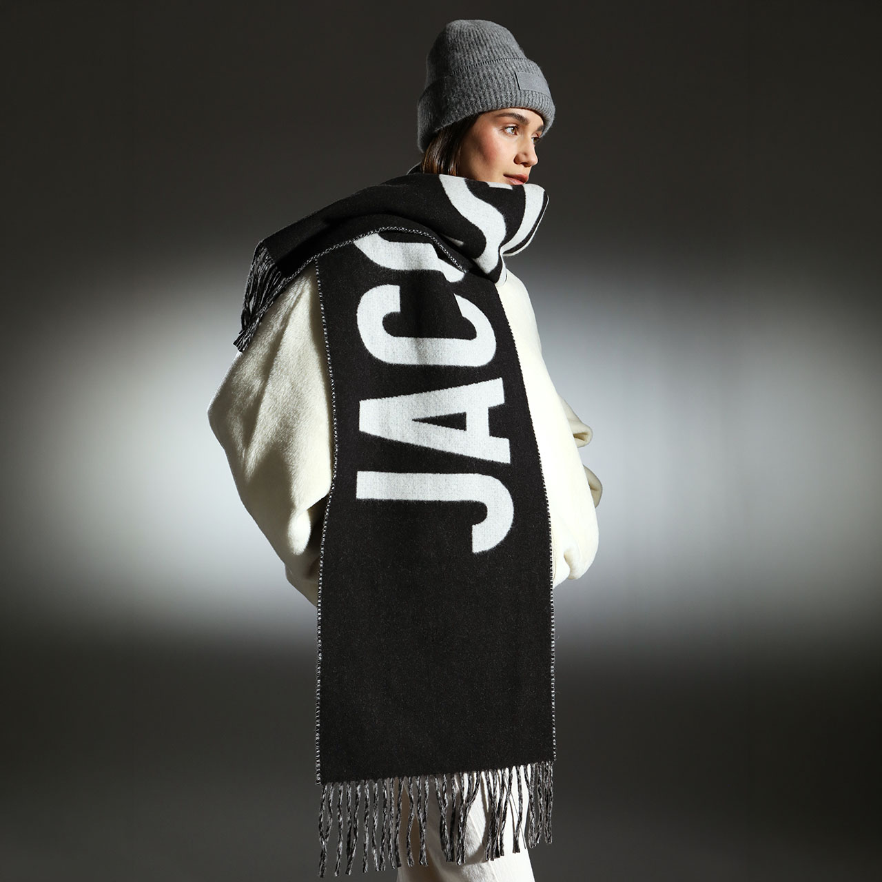 A person wearing a grey beanie and a large black and white scarf with 'JACQ' written on it. They are dressed in a voluminous white coat against a grey background.