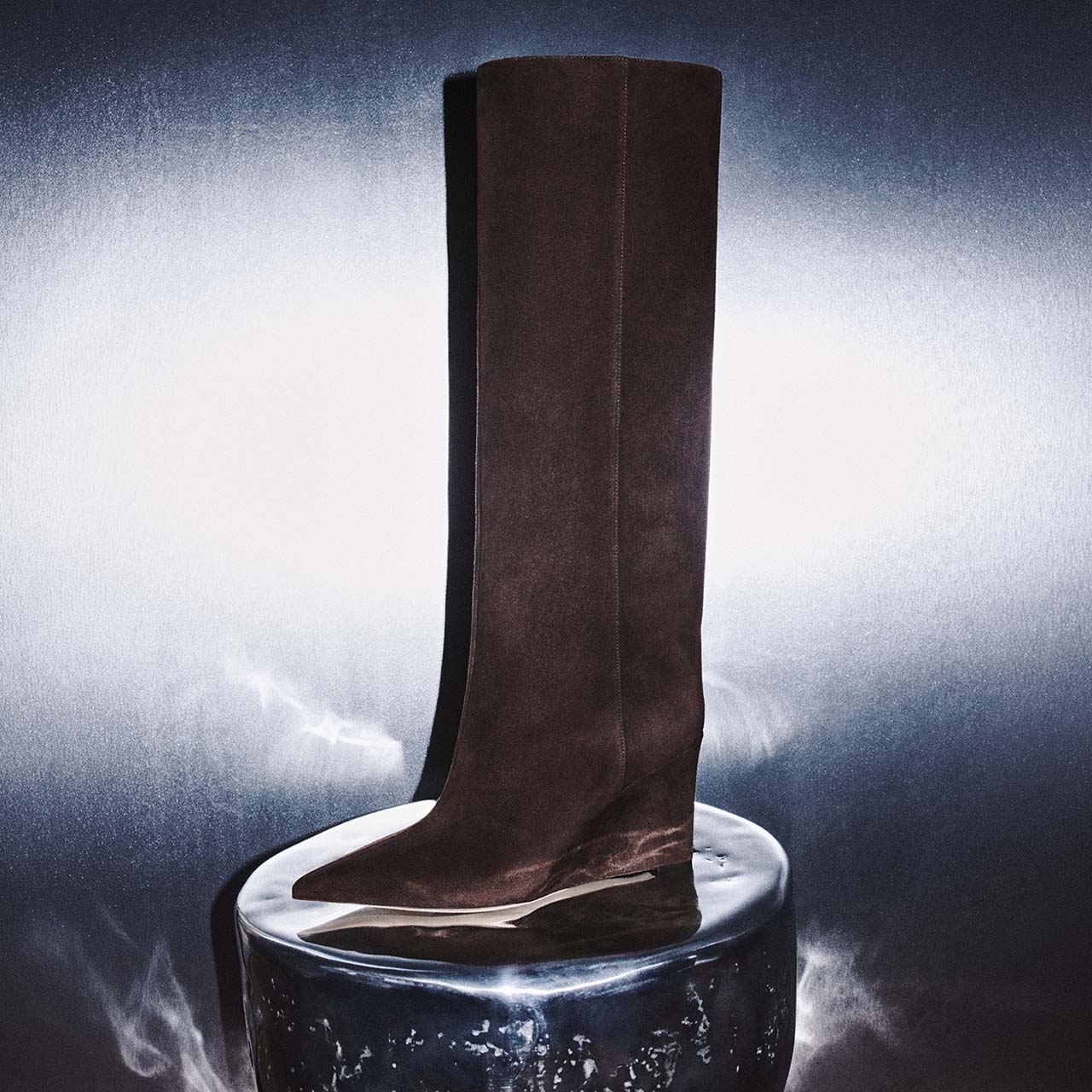 A tall, dark brown suede boot with a sleek design is placed atop a reflective, metallic surface. The background is a soft, illuminated gradient, highlighting the boot's elegant silhouette.