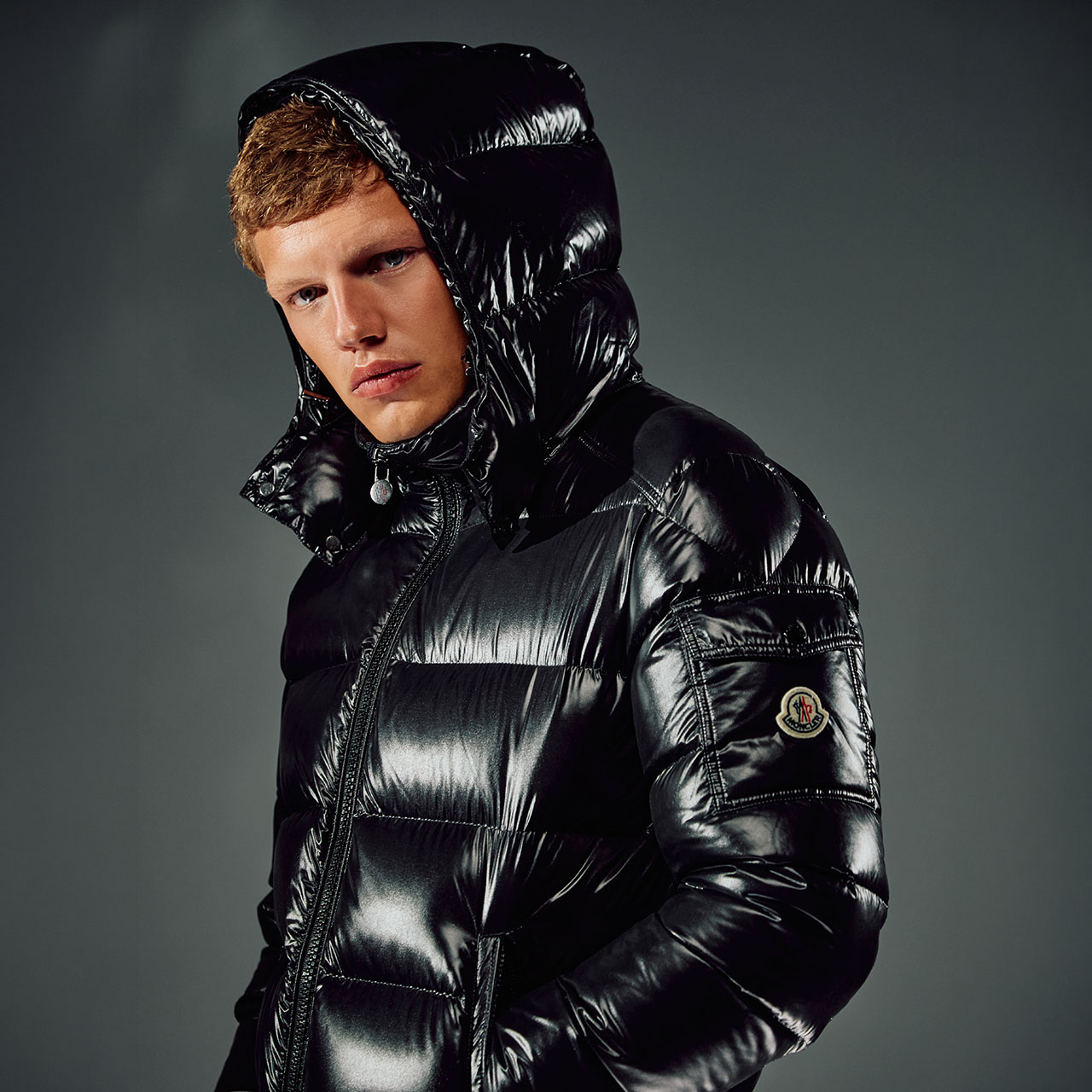Person wearing a shiny black puffer jacket with a hood, looking to the side against a dark background. The jacket has a logo on the sleeve.