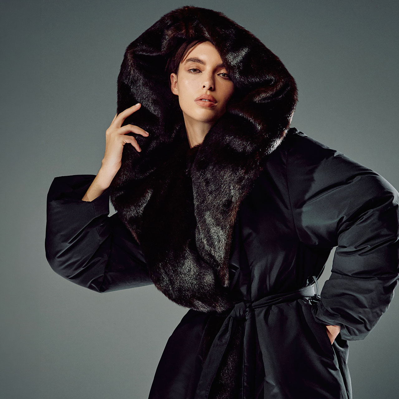 A person poses wearing a large black coat with a voluminous fur-lined hood. They have one hand raised near their face and an expression of calm confidence. The background is a simple gradient, highlighting the outfit.