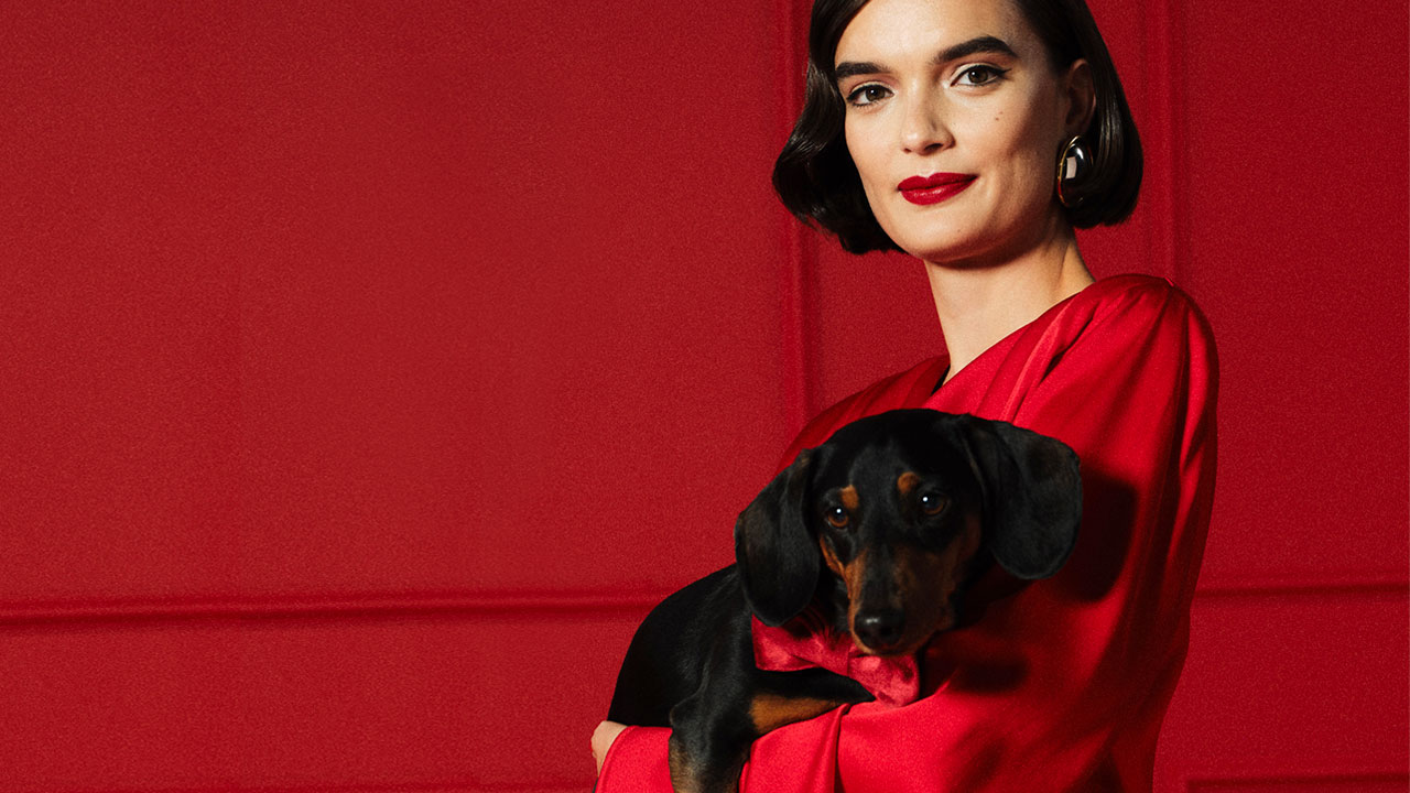 A person with long blonde hair, wearing a dark oversized coat, poses in front of a tapestry with floral and landscape patterns.