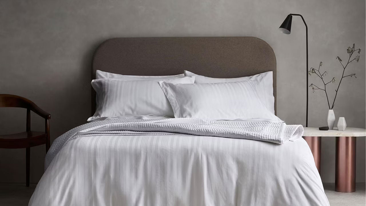 A neatly made bed with white bedding, a brown headboard, a side table with a lamp, and minimalist decor against a gray wall.