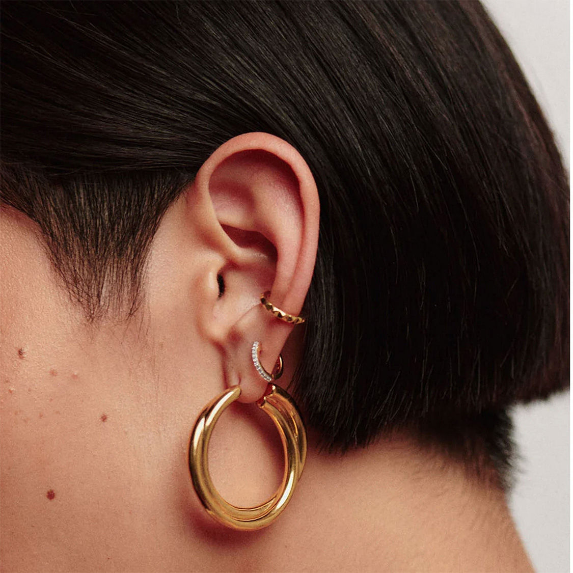 Close-up of an ear with a short black hairstyle, wearing gold hoop earrings— a larger hoop below a smaller one with a stone detail.
