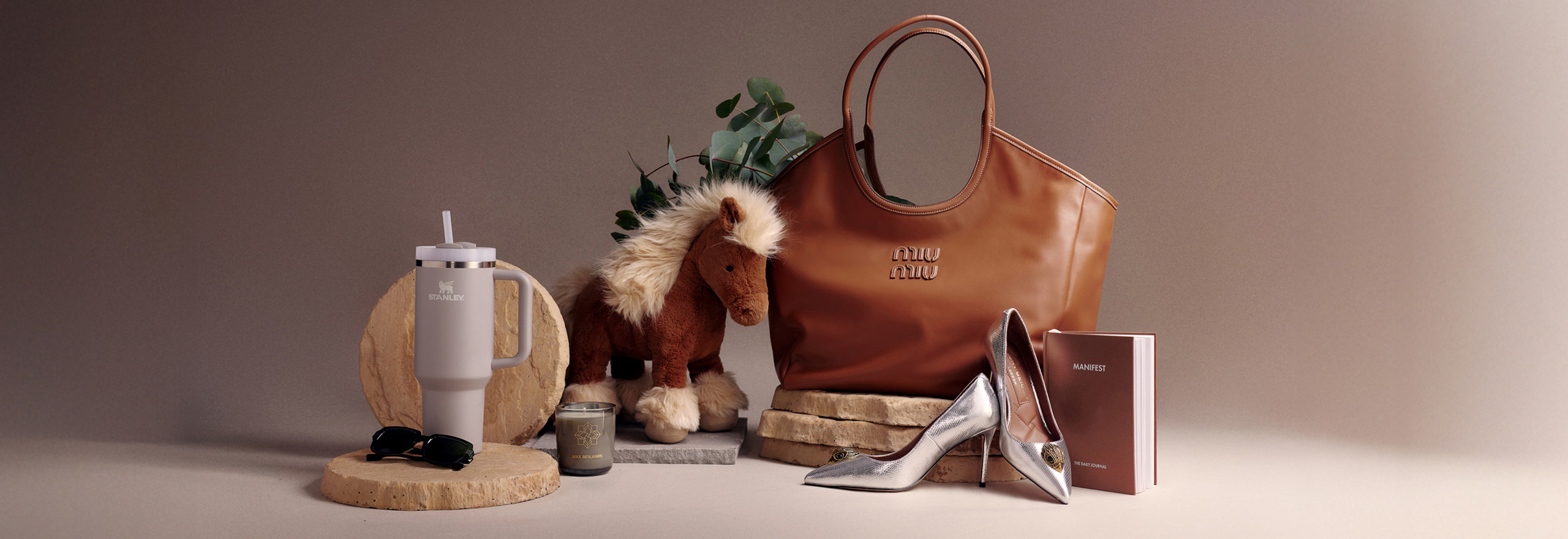 A collection of items including a brown handbag, plush horse, tumbler, sunglasses, silver shoes, candle, and a small book arranged aesthetically.