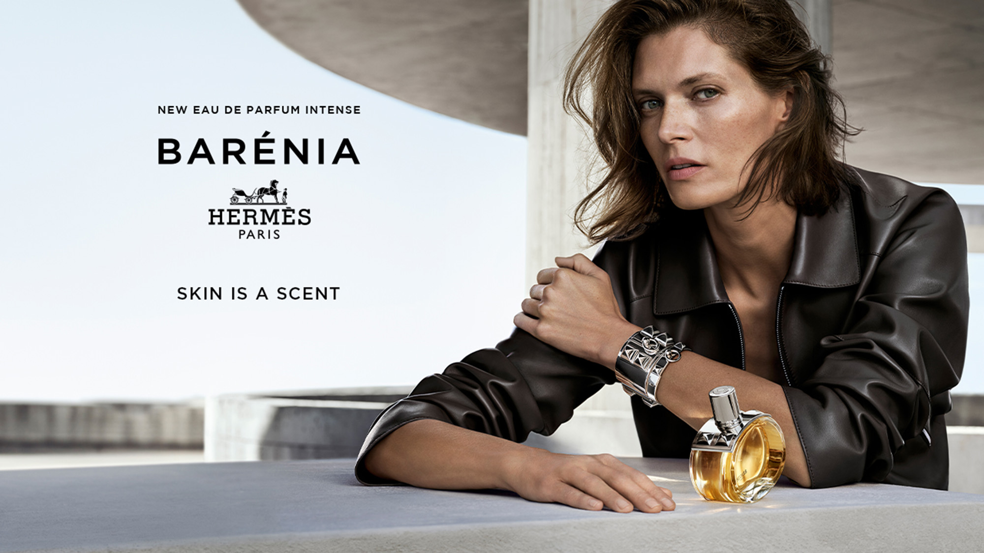A person seated at a table with a bottle of Barénia by Hermès perfume, set against a modern architectural background. Text reads 'Skin is a scent.'