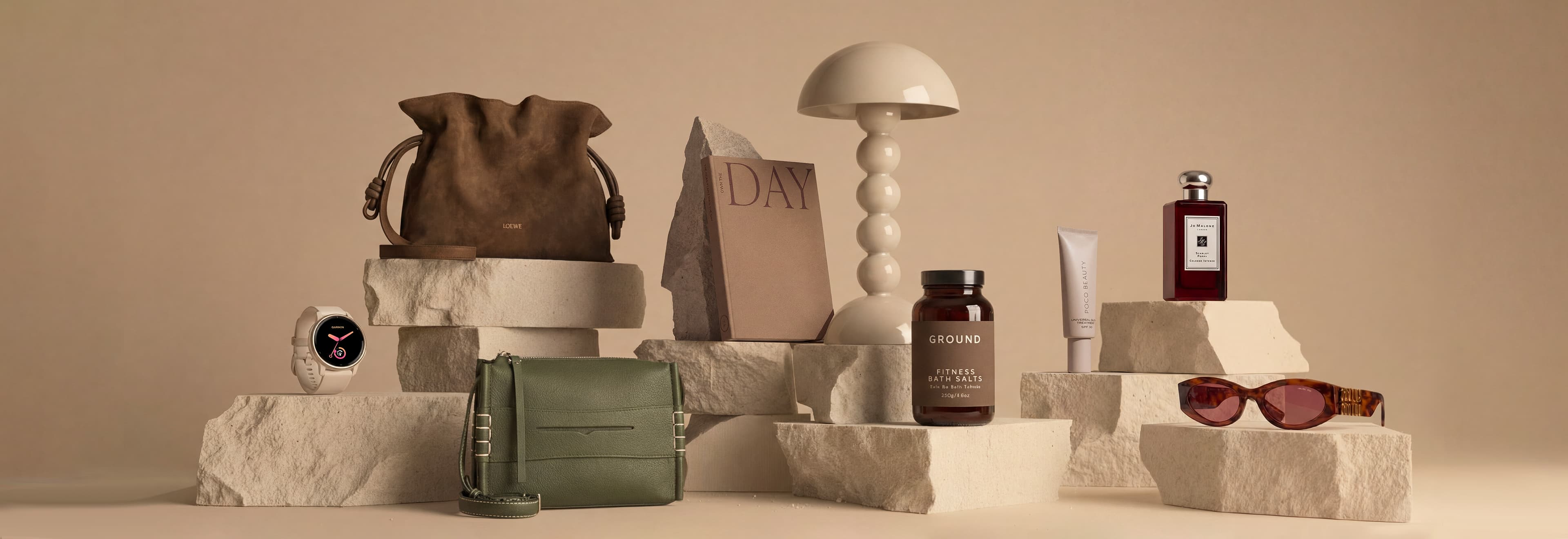 A variety of lifestyle and fashion items displayed on stone-like blocks, including bags, a lamp, a book, sunglasses, and beauty products.