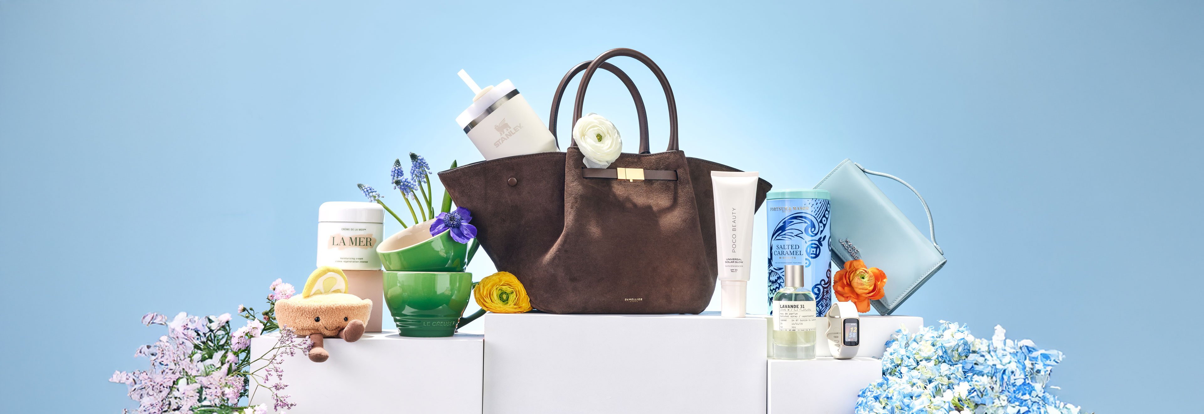 A Mother’s Day gift arrangement featuring a brown leather handbag surrounded by beauty products, candles, flowers, and small accessories displayed on white plinths against a blue background.