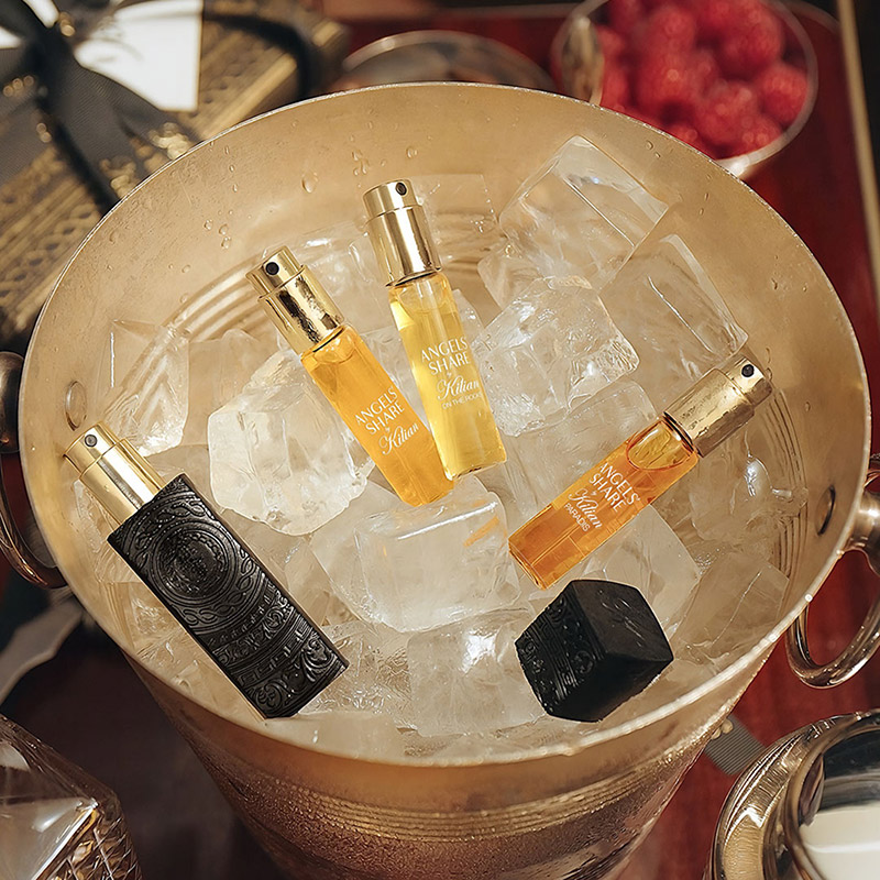 Perfume bottles chilling in an ice-filled metallic bucket. Three bottles have golden caps and different coloured liquids, while one is black with ornate details. In the background, a bowl of raspberries is partially visible.