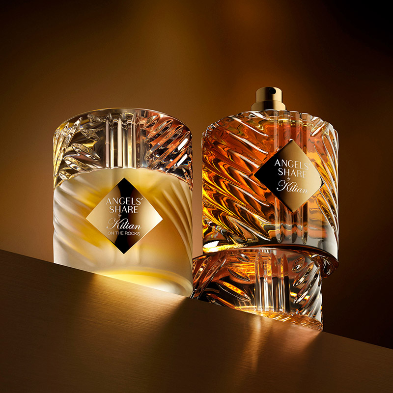 Two perfume bottles with intricate, transparent designs sit on a ledge, illuminated by warm light. Both feature black labels with 'ANGEL'S SHARE' by Kilian, enhancing their luxurious presentation.