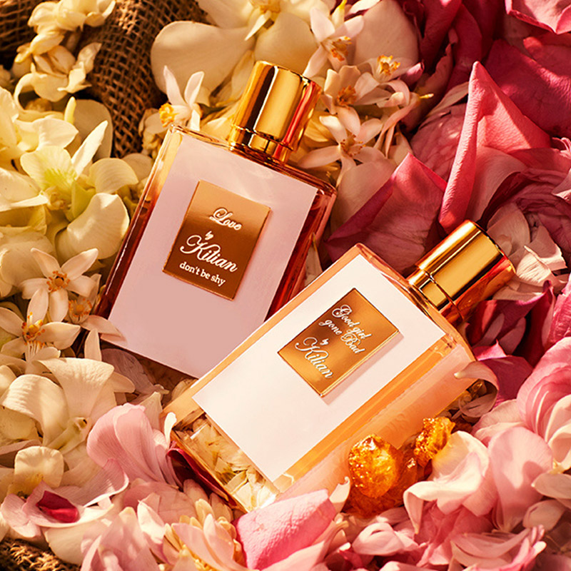 Two luxury perfume bottles with gold caps, labelled 'Love by Kilian' and 'Good Girl Gone Bad by Kilian,' surrounded by white and pink flower petals, creating an elegant and fragrant ambiance.