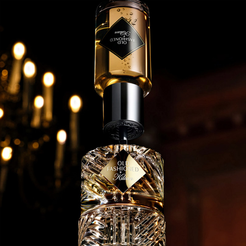 A luxurious perfume bottle labelled 'Old Fashioned' with a gold and black design is elegantly spotlighted. The background is softly blurred, featuring a dimly lit chandelier with several glowing candles, adding a sophisticated ambiance.