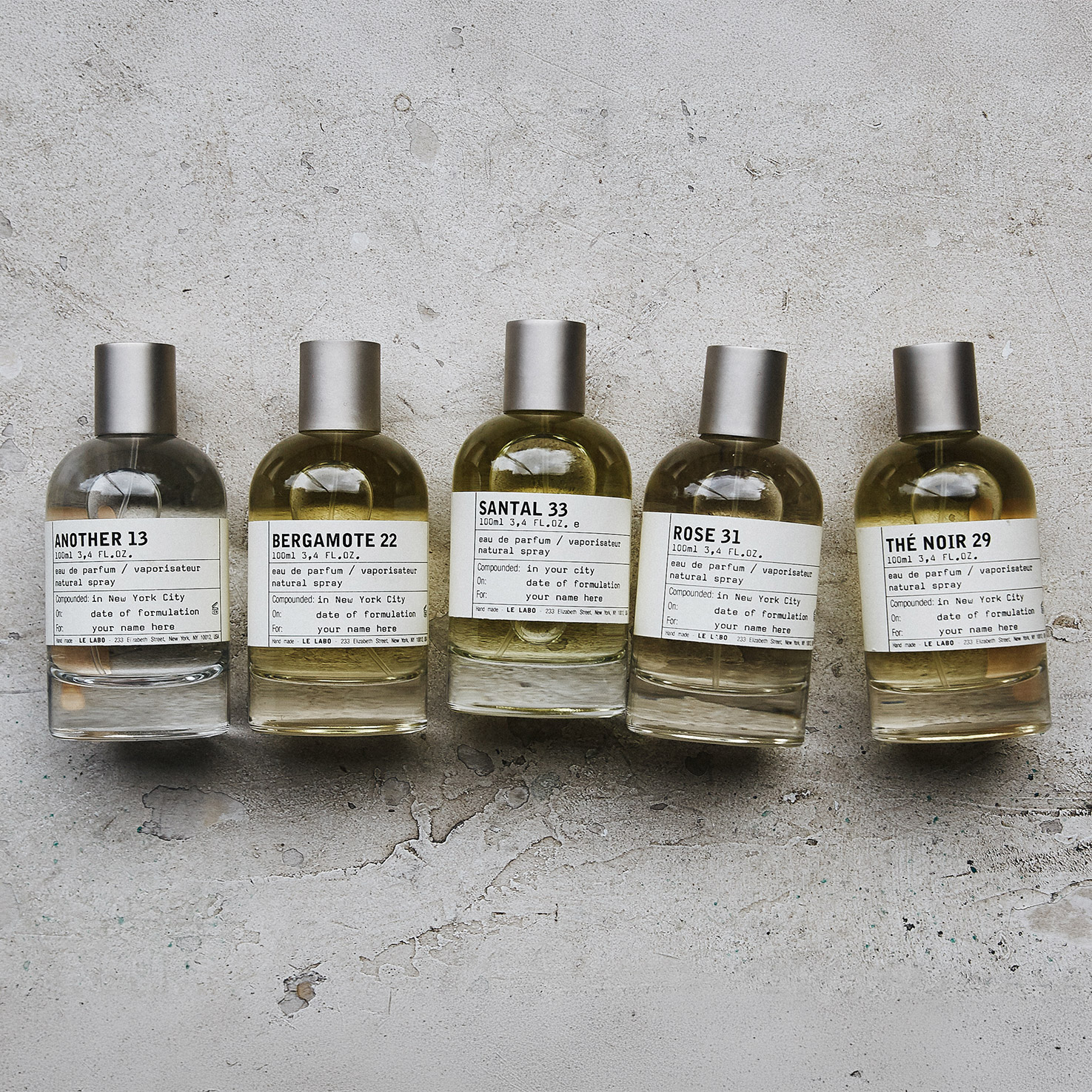 Five perfume bottles with silver caps are arranged in a row on a textured surface. Each bottle has a white label displaying the name and number of the scent: Another 13, Bergamote 22, Santal 33, Rose 31, and Th&eacute; Noir 29. The bottles contain liquids of varying hues.