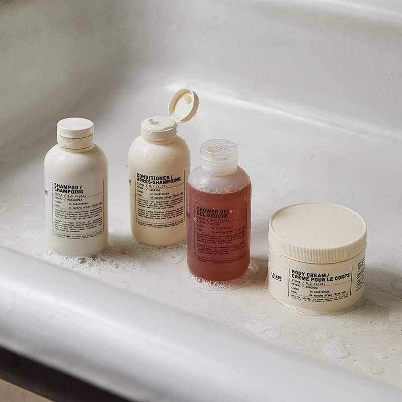 Four skincare bottles, including shampoo, conditioner, shower gel, and a body cream tub, are placed on the edge of a white bathtub. The products have minimalistic labels with text. The tub and bottles have some foam and water droplets, suggesting recent use.
