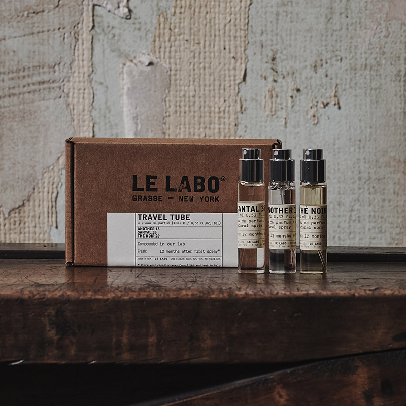 Four small perfume vials labelled 'Santal 33,' 'Another 13,' and 'Th&eacute; Noir 29' stand in front of a cardboard box marked 'Le Labo' with 'Travel Tube' written below. The box is placed on a rustic wooden surface with a textured wall in the background.