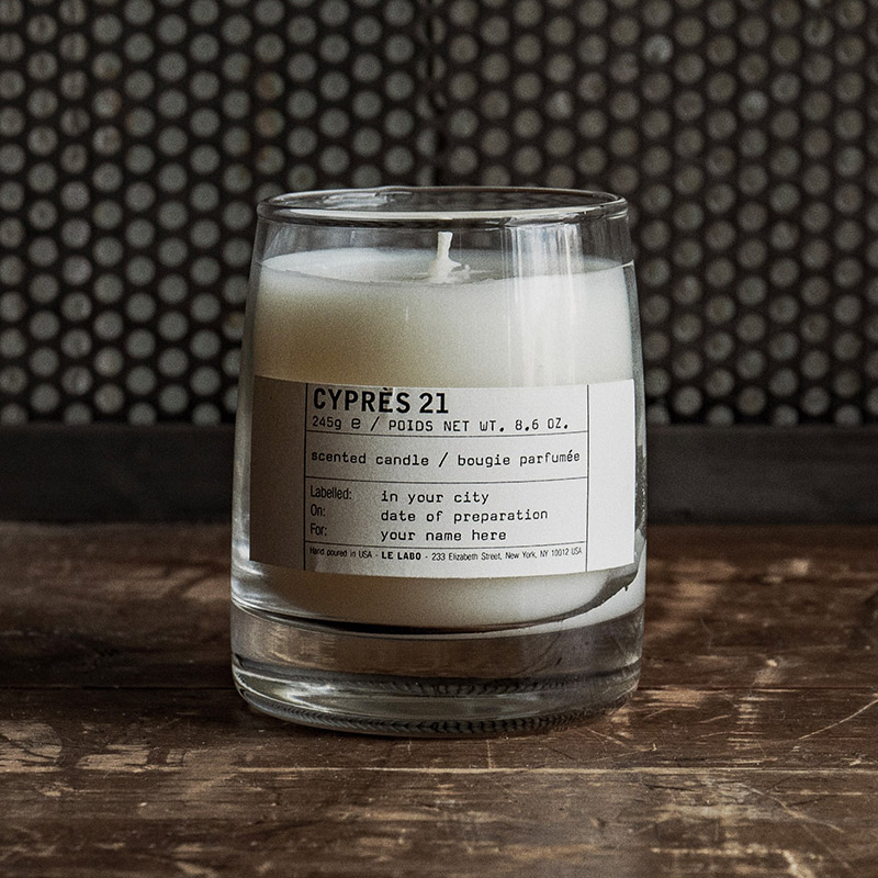 Scented candle in a clear glass holder, labelled 'CYPR&Egrave;S 21' with weight 245g/8.6 oz. The label also shows space for location, date, and name. The candle sits on a rustic wooden surface against a metal mesh background.