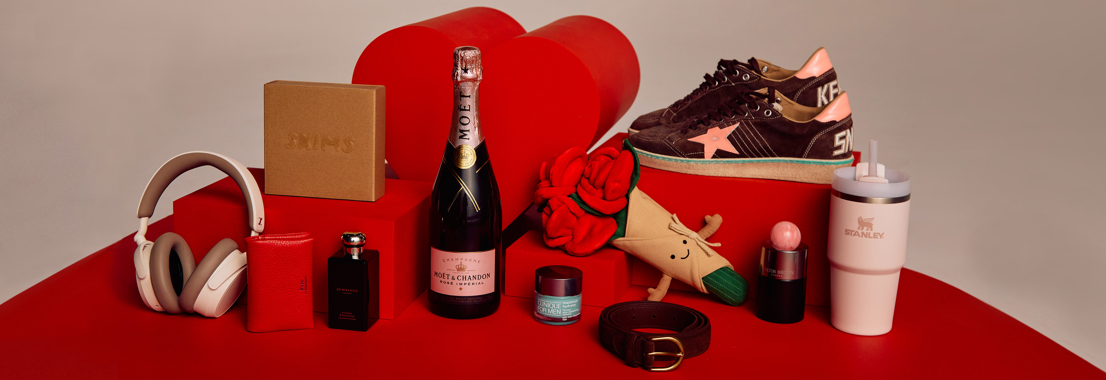 Valentine's Day-themed gifts: champagne, headphones, sneakers, cosmetics, and other items displayed on a red surface.