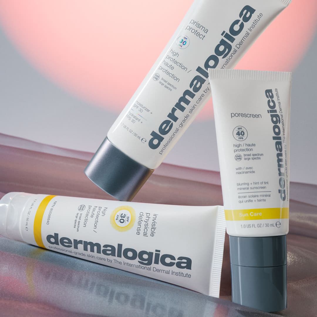 Three Dermalogica skincare products against a soft pink background. The products include a tube of Prisma Protect moisturiser, a tube of Porescreen SPF 40, and a tube of Invisible Physical Defence SPF 30. Each tube is vertically oriented, displaying the brand name prominently.