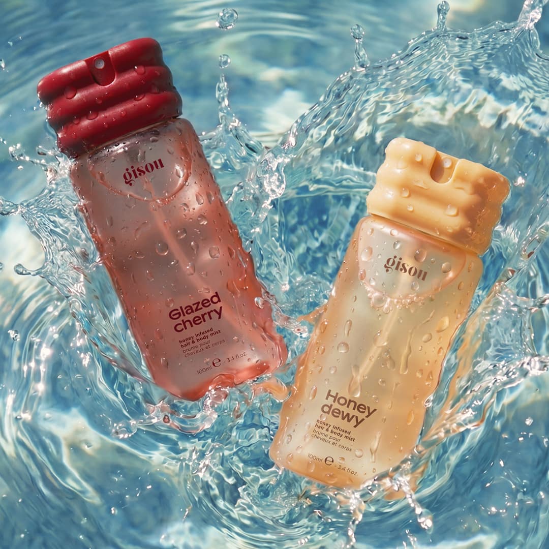 Two perfume bottles, labelled 'Glazed Cherry' and 'Honey Dewy,' are splashed with water in a dynamic, sunlit setting. The cherry bottle has a deep red cap, while the honey one is cream-coloured. Background shows rippling water.