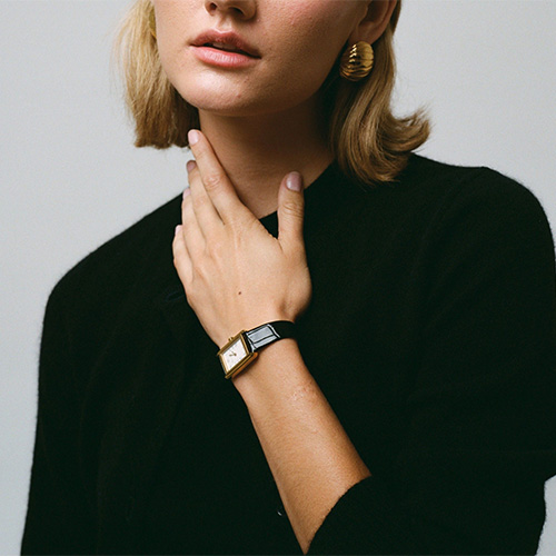 A person with short blonde hair wearing a black top, gold hoop earrings, and a watch, resting a hand on their neck.