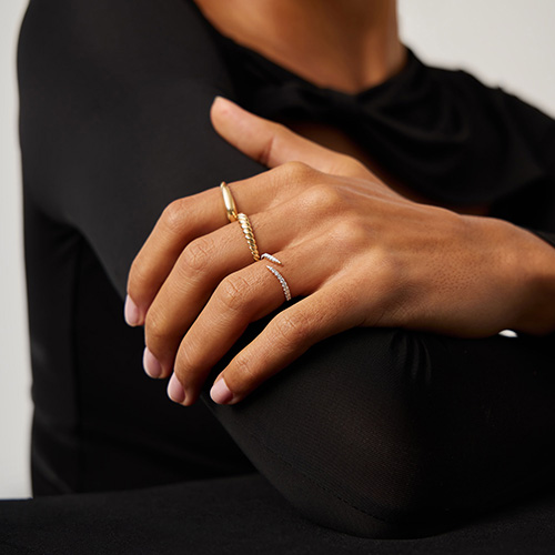 A person wearing gold and silver rings on two fingers rests their hand on a bent arm, wearing a black long-sleeve outfit.