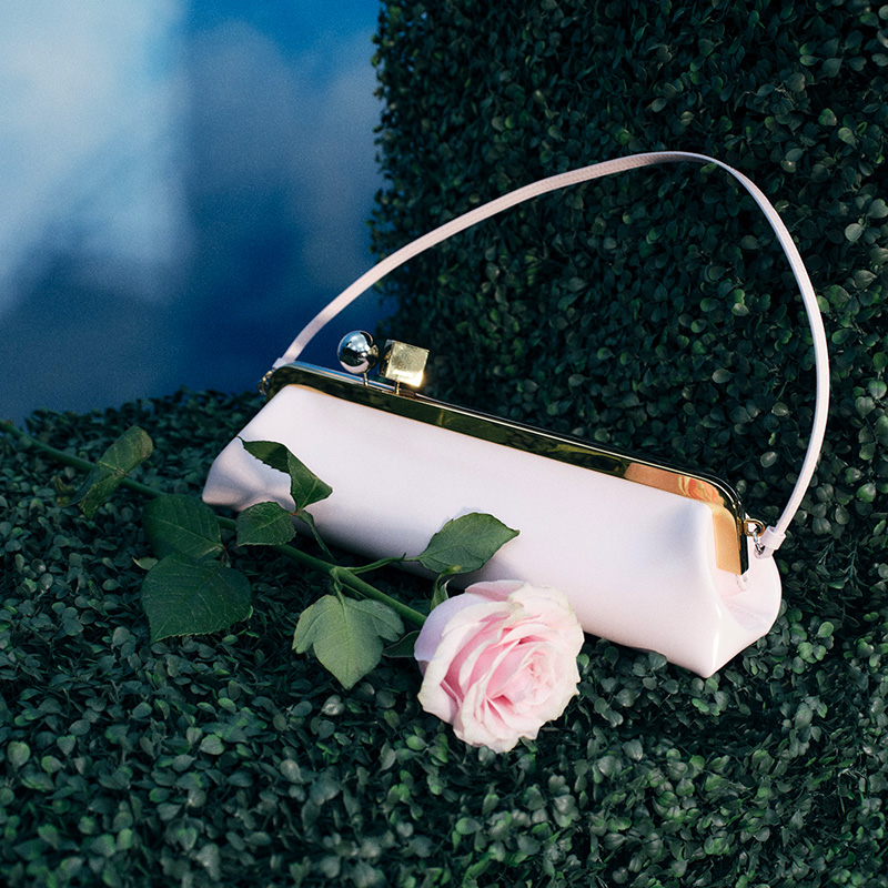 A small white handbag with a gold clasp and handle rests on green foliage, adorned with a single pink rose.
