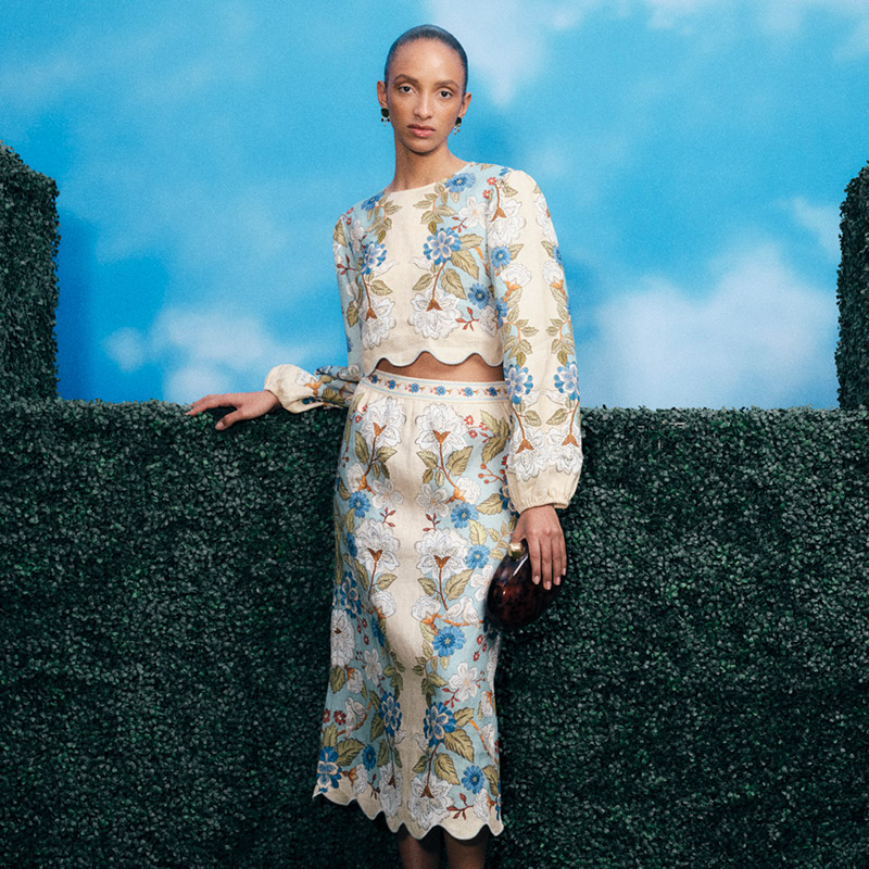 Person in a floral outfit stands against a green hedge with a blue sky backdrop.