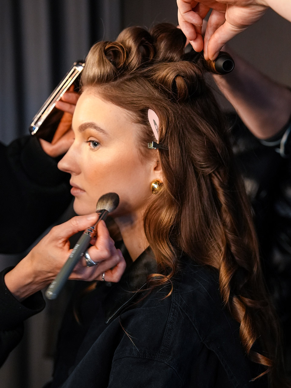 A woman having her makeup and hair done, with brushes and styling tools used by multiple hands.