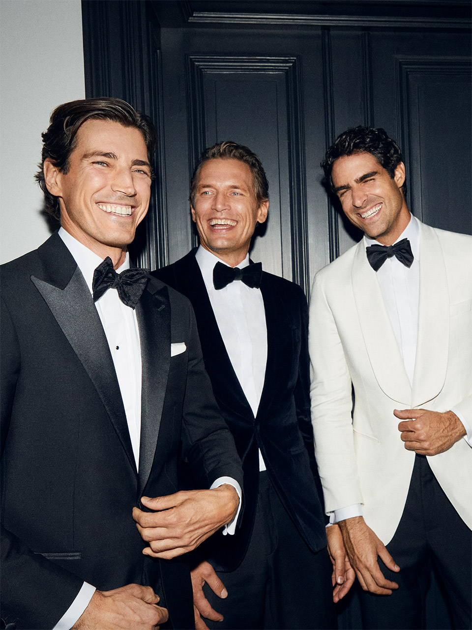Three men in tuxedos standing together, smiling and laughing.
