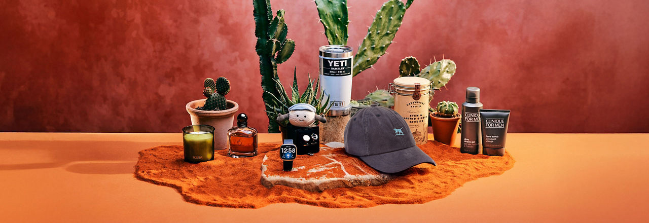 A hat, skincare products, a candle, and cacti are arranged on a table with a reddish-brown background.