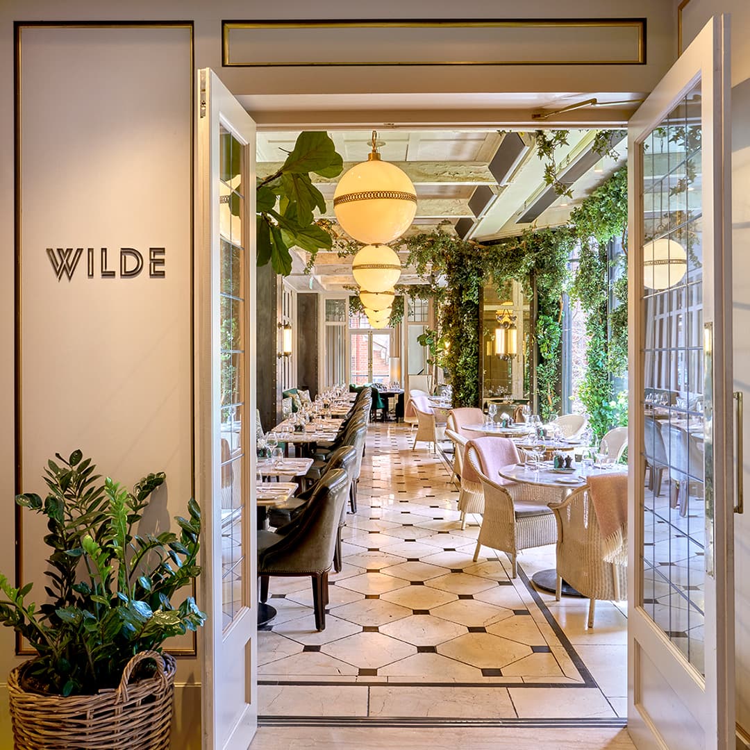 Elegant restaurant interior with greenery, stylish lighting, and neatly arranged tables. Light floods the space through open doors.