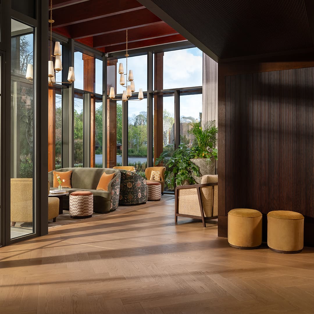 Modern lounge with large windows, wooden floors, plush seating, and hanging lights. Lush greenery is visible outside.