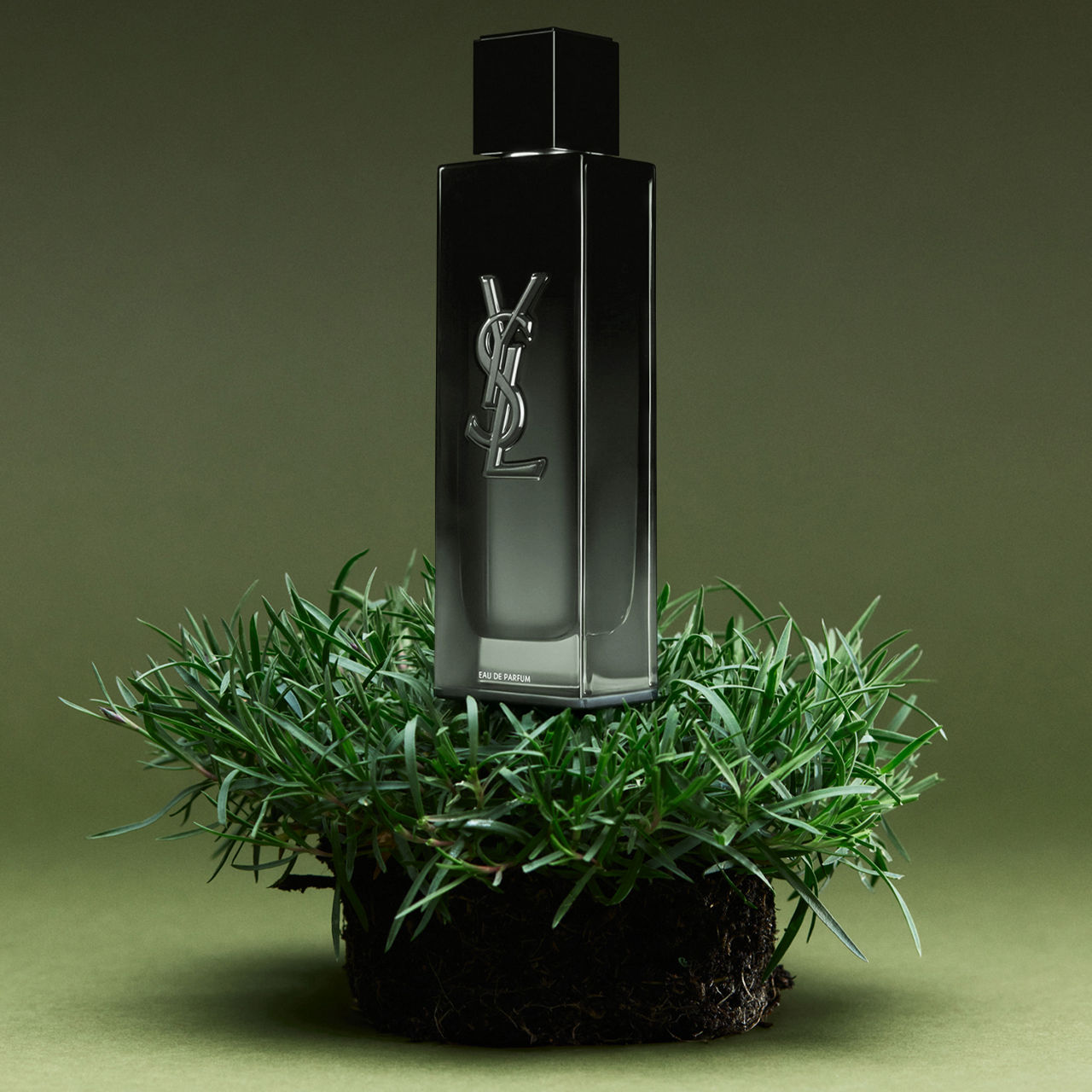 A sleek, dark perfume bottle with a vertical logo, placed atop a small mound of lush green grass against a green background.