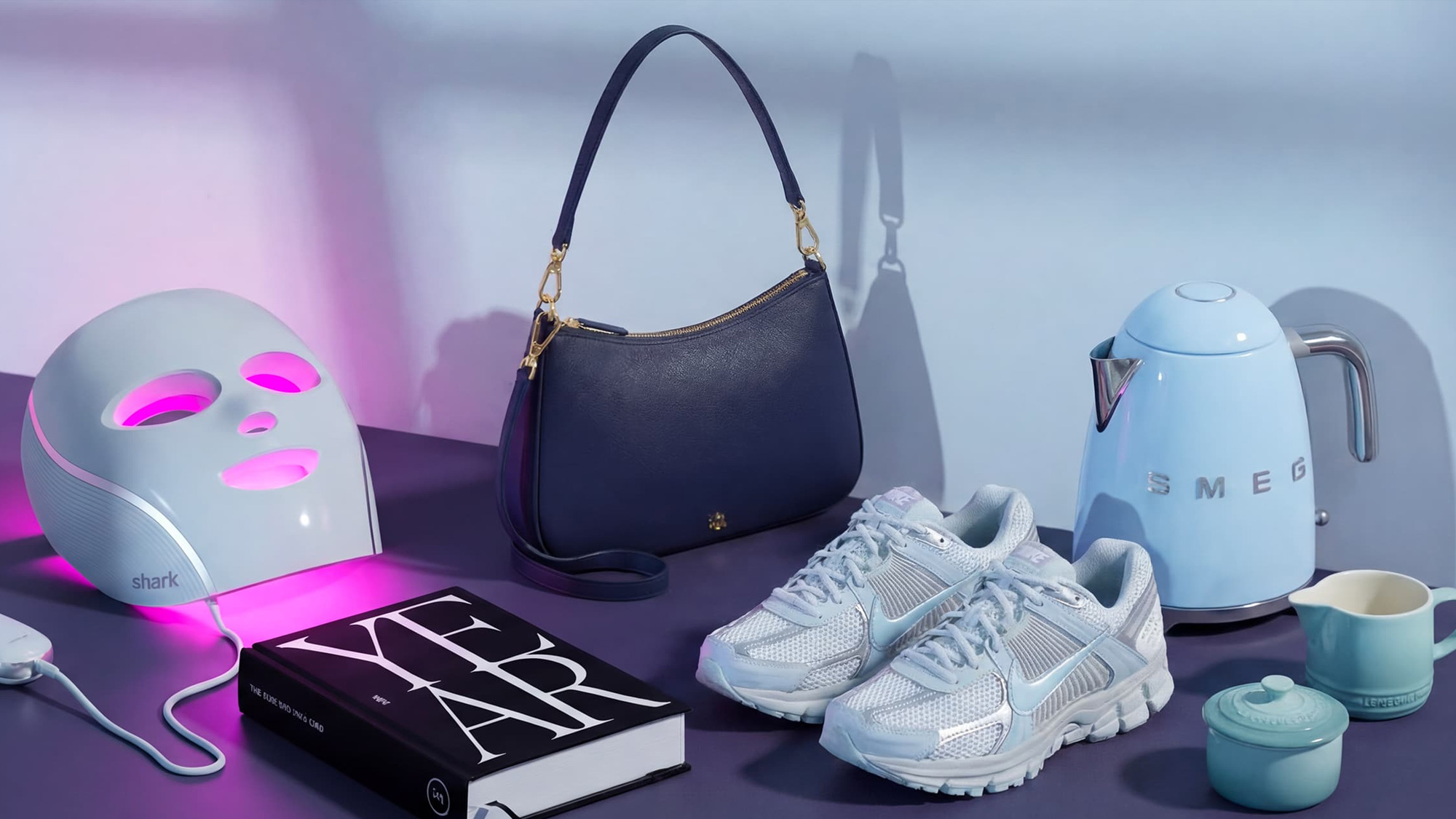Assorted items including a face mask, handbag, sneakers, book, kettle, and cups displayed on a purple surface with a pink and blue background.