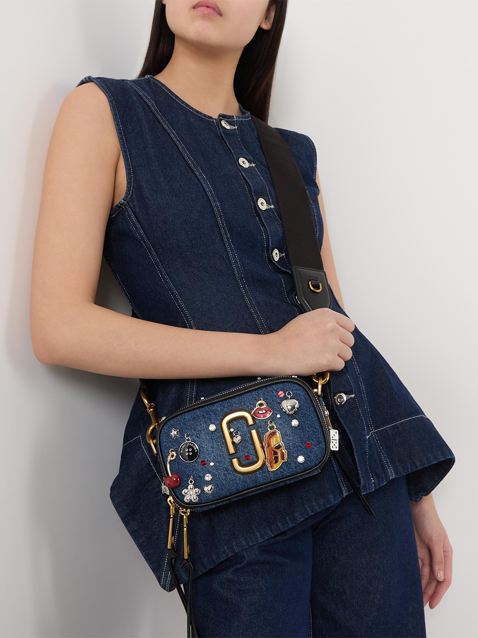 A person wearing a sleeveless denim top and matching denim trousers is holding a small blue handbag adorned with various colourful embellishments. The handbag features a distinctive gold clasp and is slung across the body with a black strap.