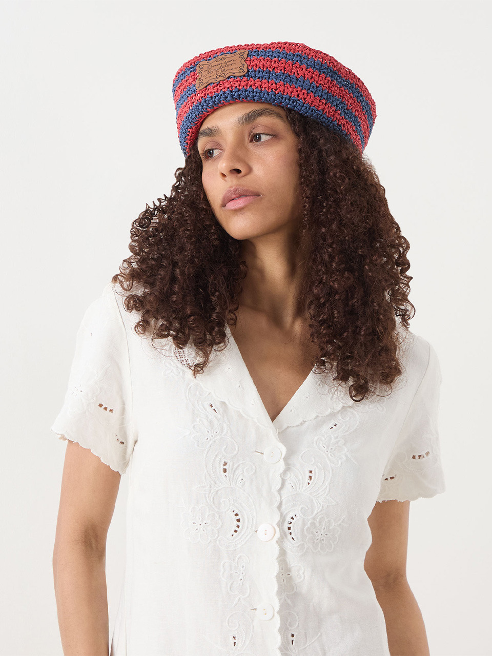A person with curly hair wearing a striped red and blue crochet beret and a white embroidered blouse. They are looking to the side against a plain background.
